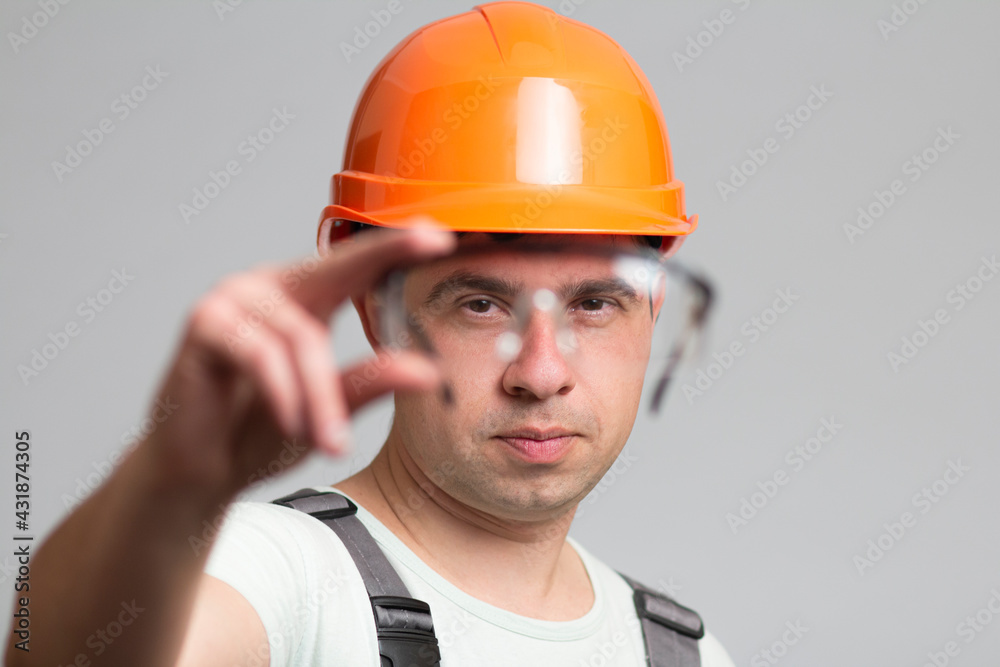 protective glasses as element of builder's uniform in hands of young man, construction worker in helmet and overalls on grey background