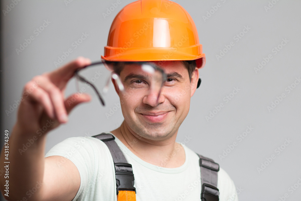 protective glasses as element of builder's uniform in hands of young man, construction worker in helmet and overalls on grey background protective glasses as element of builder's uniform in hands of young man, construction worker in helmet and overalls on grey background