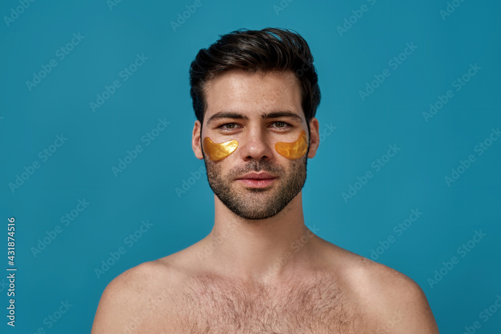 Portrait of brunette caucasian guy with golden gel under eye patches looking at camera, posing isolated over blue background Portrait of brunette caucasian guy with golden gel under eye patches looking at camera, posing isolated over blue background
