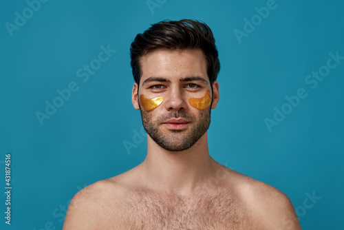 Portrait of brunette caucasian guy with golden gel under eye patches looking at camera, posing isolated over blue background