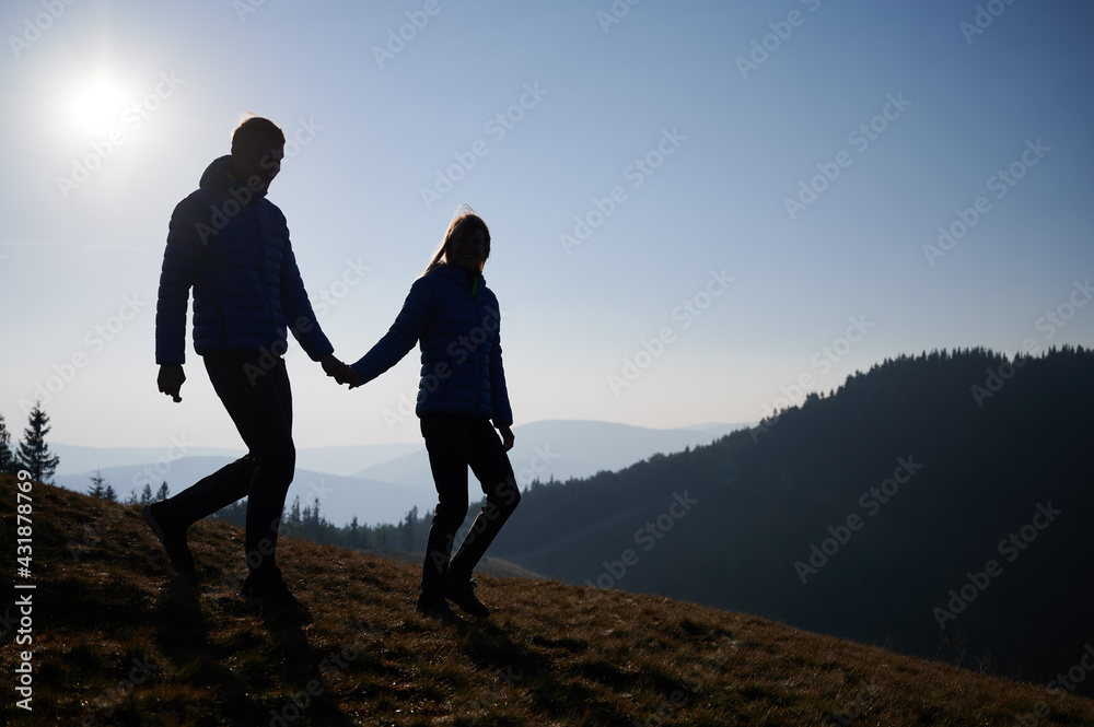 Full length of happy couple in love holding hands and smiling while walking down grassy hill early in the morning. Silhouettes of young man and woman travelers spending time together in mountains.