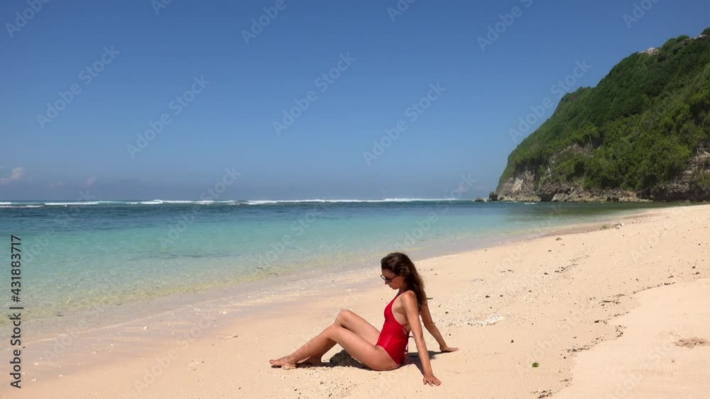 Tanned slim brunette woman in red swimsuit sitting near water, enjoying, posing on wild tropic beach with blue water and cliff on background, Bali, Indonesia Tanned slim brunette woman in red swimsuit sitting near water, enjoying, posing on wild tropic beach with blue water and cliff on background, Bali, Indonesia