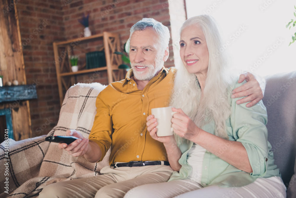 Photo of old grey haired retired pensioner couple sit couch drink tea watch tv indoors inside house home - obrazy, fototapety, plakaty 
