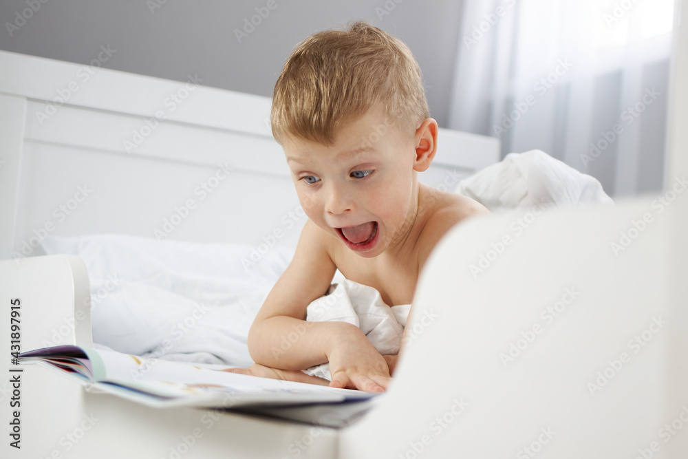 The child is surprised by what he has read in the new book. Funny little boy with a book in a white bed The child is surprised by what he has read in the new book. Funny little boy with a book in a white bed