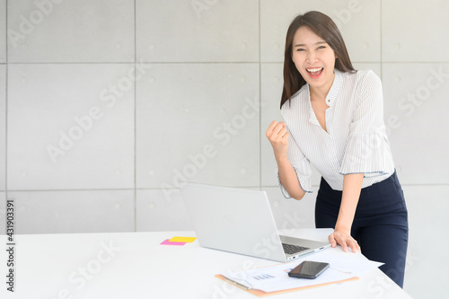 Beautiful excited Asian woman celebrate success arms up with laptop and document in office. business success concept 