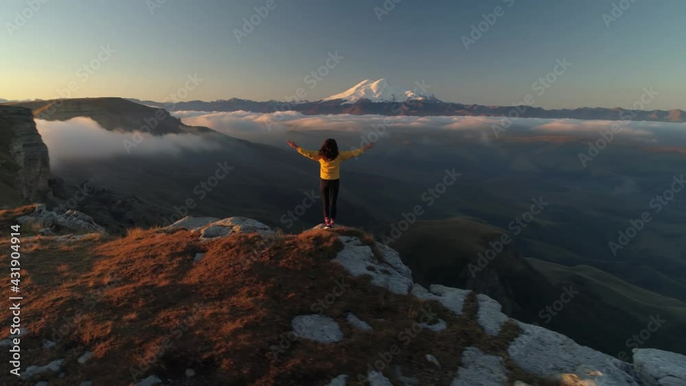 Girl hands spread stands on top of mountain looks at Mount Elbrus. Caucasus emotional inspirational achivement. Travel film Russia. Orange sunset. Scenic clouds fog below. Bermamyt plateau 4k