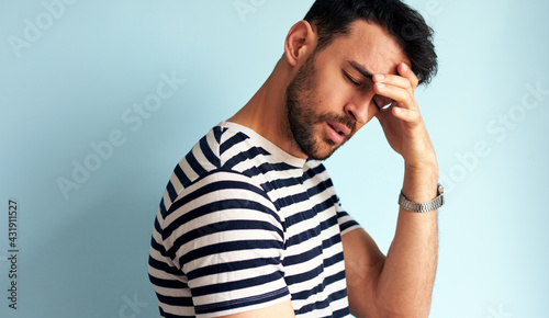 Portrait of a unhappy young man keeps hand on forehead with closed eyes, dressed in striped t-shirt, isolated over blue studio wall. A male has a headache.