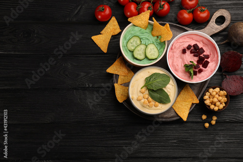 Different kinds of tasty hummus served with nachos and ingredients on black wooden table, flat lay. Space for text