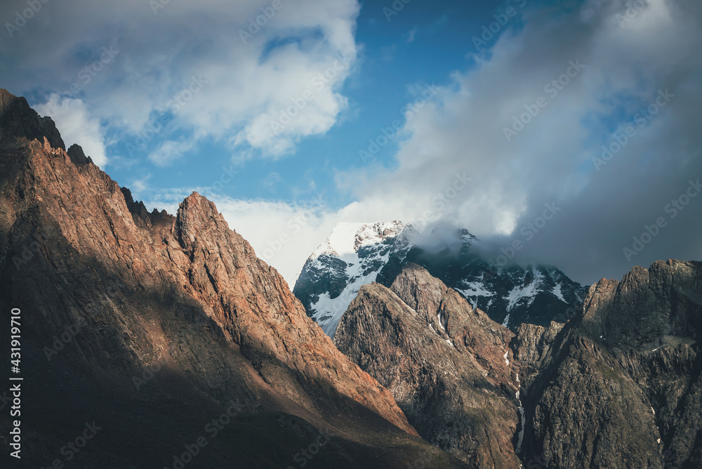 Scenic landscape with great rocks and snowy mountains in sunlight in low clouds. Wonderful view to mountain top with snow in sunshine in cloudy sky. Awesome scenery with sunlit snow-white pinnacle.