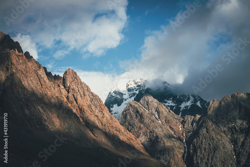 Scenic landscape with great rocks and snowy mountains in sunlight in low clouds. Wonderful view to mountain top with snow in sunshine in cloudy sky. Awesome scenery with sunlit snow-white pinnacle. © Daniil