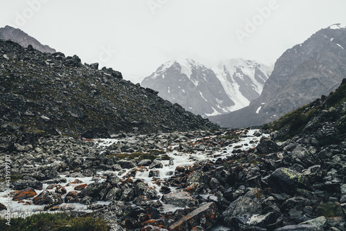 Atmospheric landscape with mountain creek among moraines in rainy weather. Bleak scenery with milky river from snowy mountains. Stones with moss and lichen in water stream. Mountain river among rocks.