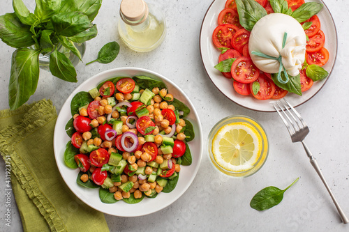 Fresh summer dinner: mediterranean salad with chickpea, tomatoes, cucumbers, spinach and caprese salad with burrata cheese, basil leaves and tomatoes. Water with lemon. Light background, copy space.