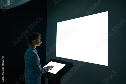 Mock up, futuristic, template and education concept. Woman using electronic kiosk and looking at white blank large interactive wall display in dark room of modern technology museum - mockup image