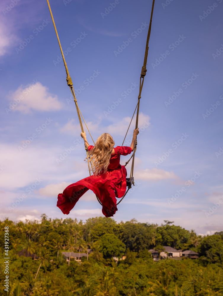 Bali swing trend. Caucasian woman in long red dress swinging in the jungle rainforest. Vacation in Asia. Travel lifestyle. Blue sky. View from back. Bongkasa, Bali, Indonesia