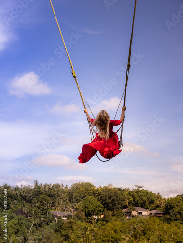 Bali swing trend. Caucasian woman in long red dress swinging in the jungle rainforest. Vacation in Asia. Travel lifestyle. Blue sky. View from back. Bongkasa, Bali, Indonesia