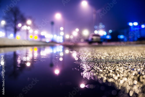 Foggy night in the big city  the street lighting lanterns and a parked car. Close up view from the level of the puddle on the pavement