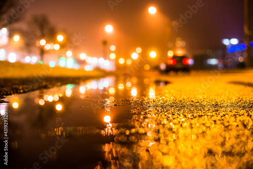Foggy night in the big city, the street lighting lanterns and riding car. Close up view from the level of the puddle on the pavement