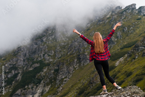 Traveler girl standing raised arms in the top of mountain. Clouds and rocks in the background. Red checkered shirt.