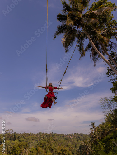 Bali swing trend. Caucasian woman in long red dress swinging in the jungle rainforest. Vacation in Asia. Travel lifestyle. Blue sky. Bongkasa, Bali, Indonesia