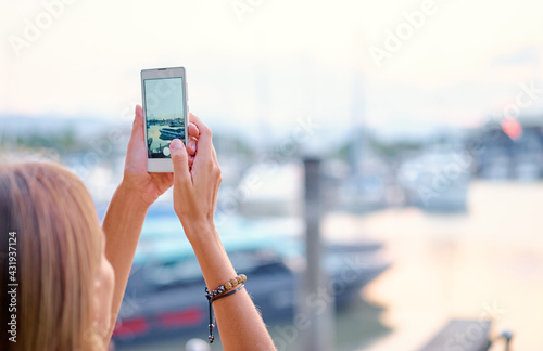 Tourism concept. Young woman taking photo on smartphone while standing on the harbor seafront.