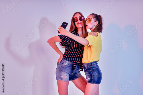 Two teenage girls in summer clothes standing in studio and making selfie on white background. Mixed light