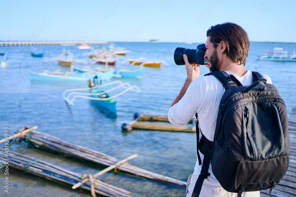 Obraz premium Photography and travel. Young man with rucksack taking photo with his camera on the sea beach near fishing boats.