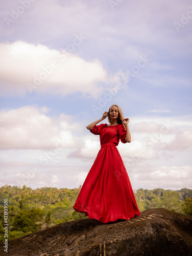 Bali trend photo. Caucasian woman in long red dress standing on big stone in tropical rainforest. Vacation in Asia. Travel lifestyle. Breathtaking view. Bongkasa, Bali, Indonesia © Olga