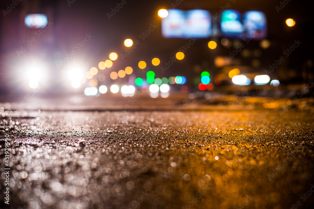 Rainy night in the big city, glare from the headlights of the parked car and a passing near the stream of cars. Close up view from the sidewalk level