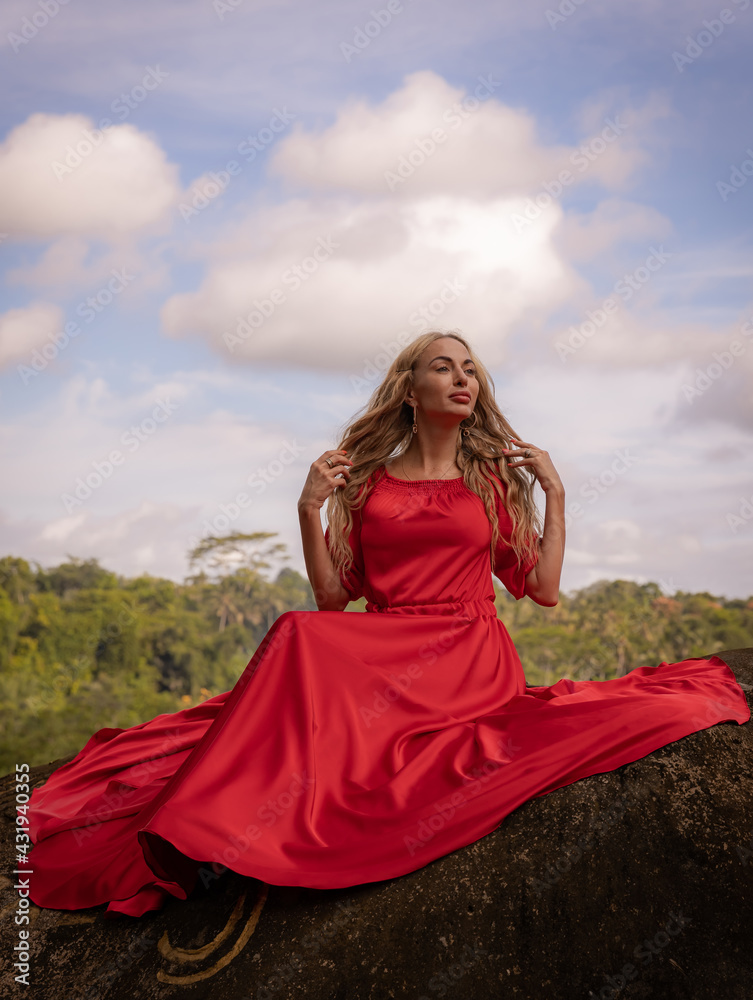 Bali trend photo. Caucasian woman in long red dress sitting on big stone in tropical rainforest. Vacation in Asia. Travel lifestyle. Breathtaking view. Bongkasa, Bali, Indonesia