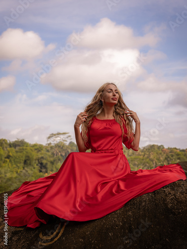 Bali trend photo. Caucasian woman in long red dress sitting on big stone in tropical rainforest. Vacation in Asia. Travel lifestyle. Breathtaking view. Bongkasa  Bali  Indonesia