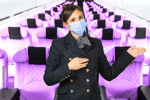 A flight attendant with a surgical mask points with her arms in the aisle of an airplane. Subject on a blurred background.