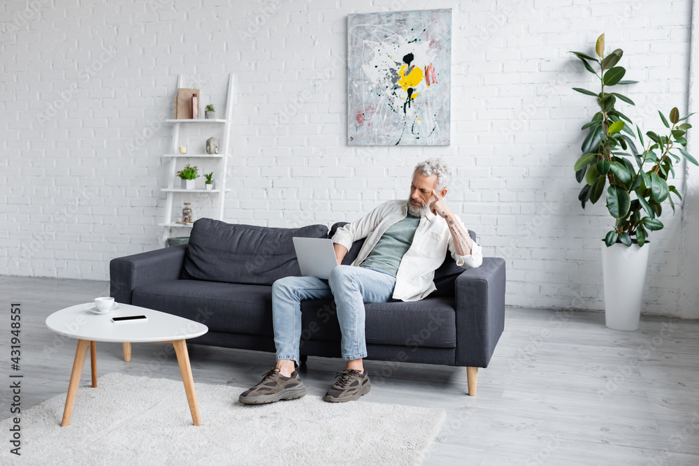 man with grey hair using laptop while sitting on couch near cup of coffee and smartphone with blank screen on coffee table. - obrazy, fototapety, plakaty 