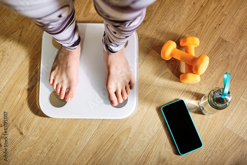 Woman checking weight while standing on weight scale by dumbbell, water bottle and mobile phone at home