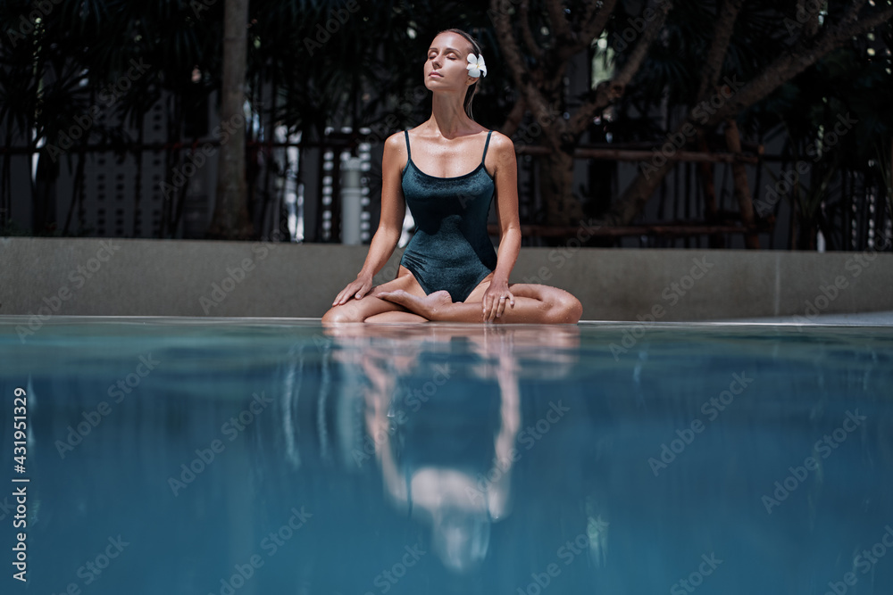 Wellness spa, vacation and yoga meditation concept. Pretty young woman in swimsuit sitting in lotus position near the swimming pool.