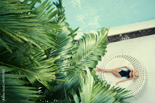 Enjoying suntan. Vacation concept. Top view of slim young woman in swimwear on the sun lounger near the swimming pool with palm trees.