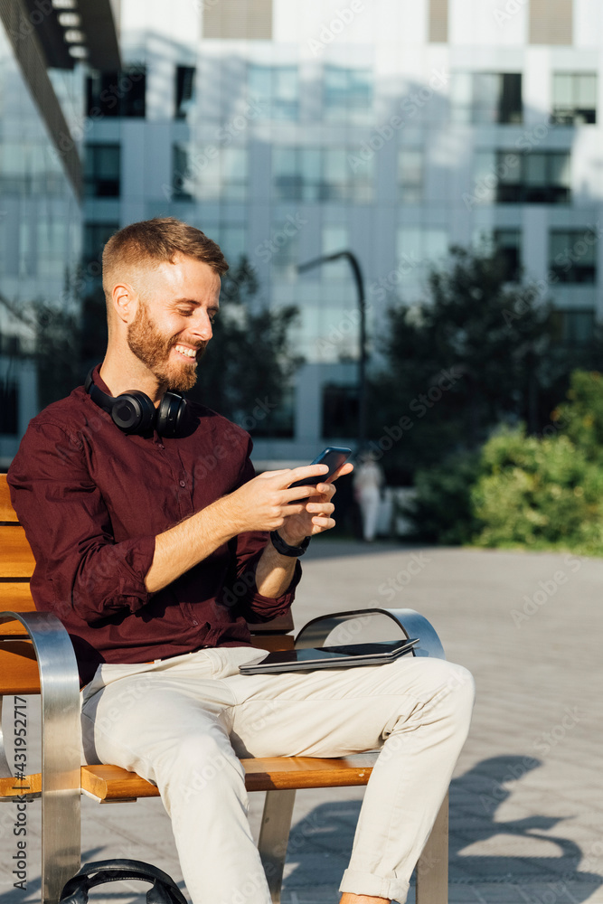 Smiling businessman using smart phone while sitting on bench during sunny day Smiling businessman using smart phone while sitting on bench during sunny day