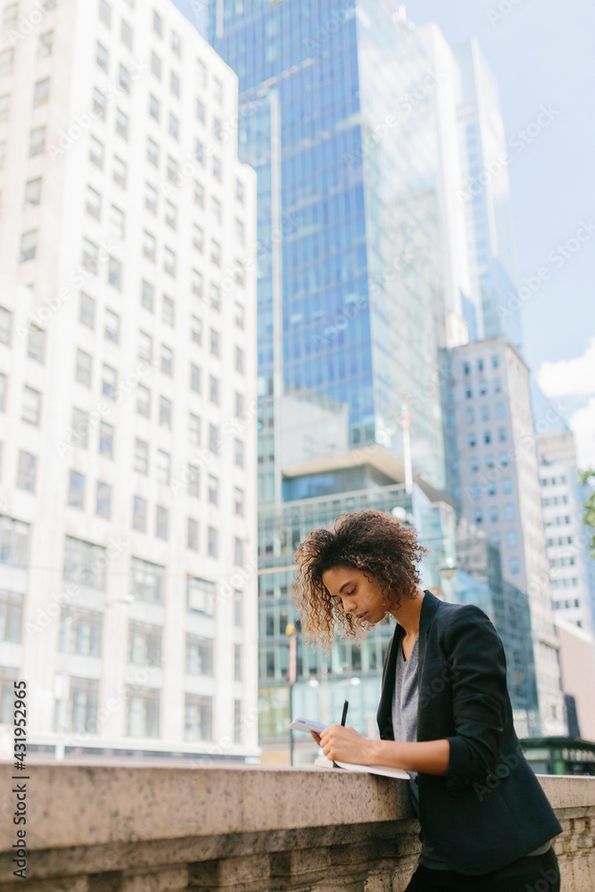 Afro female professional writing while leaning on retaining wall near office buildings Afro female professional writing while leaning on retaining wall near office buildings