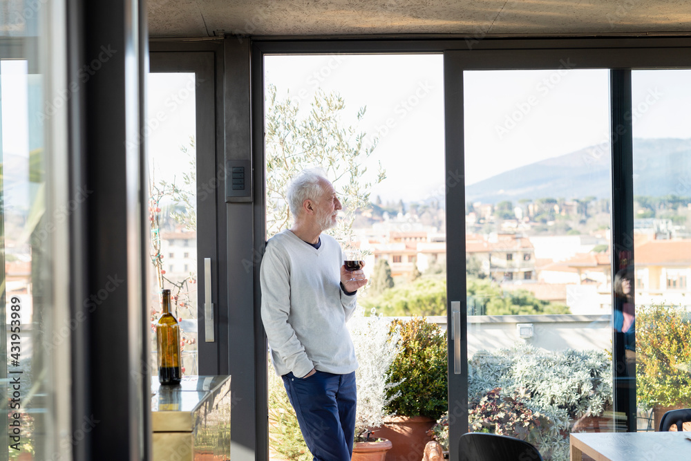 Senior man holding wineglass while leaning by sliding door at home Senior man holding wineglass while leaning by sliding door at home