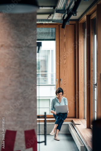 Female entrepreneur working on laptop while sitting over alcove window seat at office