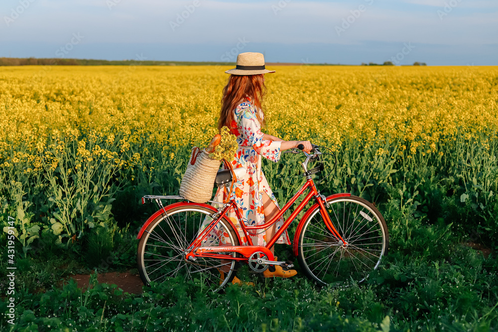 Fototapeta premium Girl with a red retro bicycle going along the yellow field of flowers.