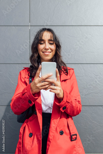 Smiling young businesswoman using phone while standing against gray color wall photo