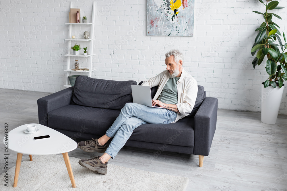 bearded man with grey hair sitting on couch and using laptop in living room. - obrazy, fototapety, plakaty 