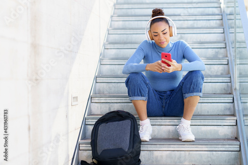 Young woman with backpack and headphones using mobile phone while sitting on steps photo