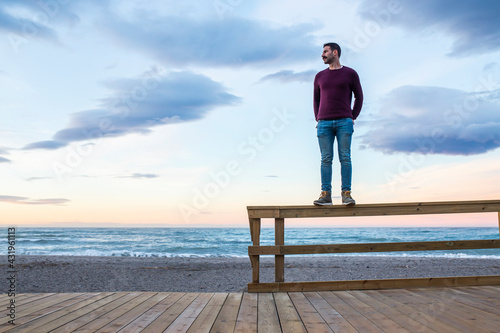 Man with hands in pockets standing on wooden railing at beach against sky