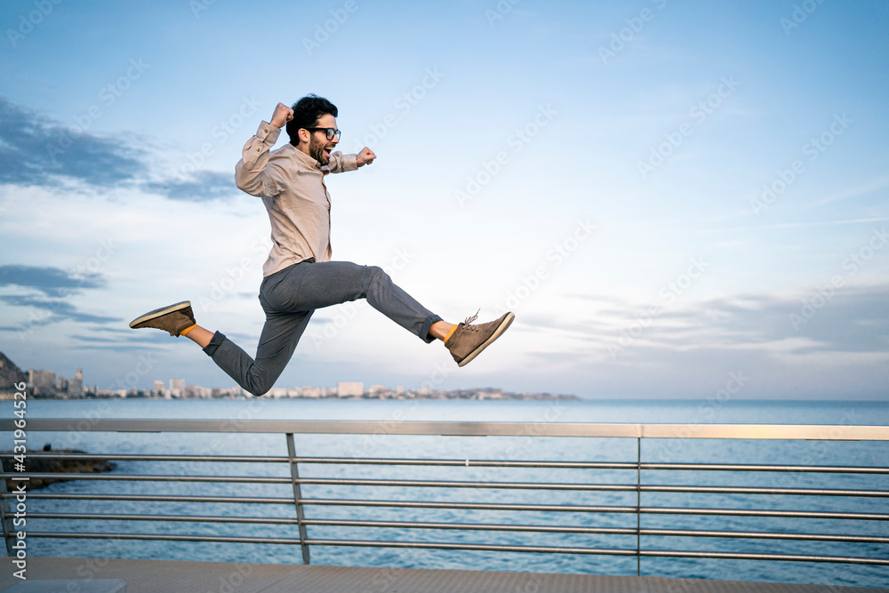 Carefree male entrepreneur with arms raised jumping by railing in front of blue sky Carefree male entrepreneur with arms raised jumping by railing in front of blue sky