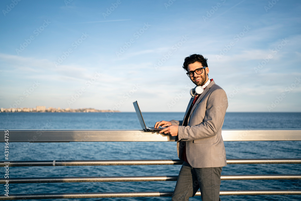 Smiling male business professional with laptop standing at railing by sea Smiling male business professional with laptop standing at railing by sea