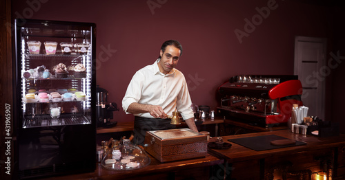 Young cheerful barista wearing black apron and white shirt while preparing turkish coffee in a modern coffee shop