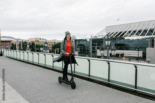 Happy woman riding electric push scooter on footpath near railing in city