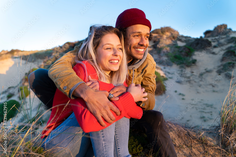 © steve brookland/Westend61 - Man wearing knit hat embracing beautiful woman while sitting on sand dune © steve brookland/Westend61 - Man wearing knit hat embracing beautiful woman while sitting on sand dune