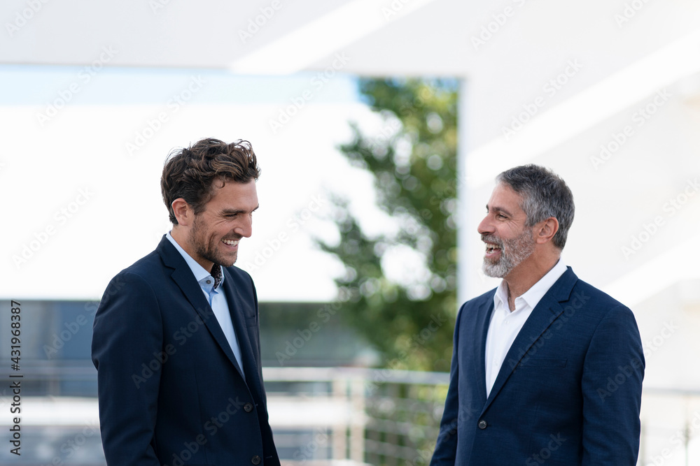 Businessmen laughing while standing at office building terrace Businessmen laughing while standing at office building terrace